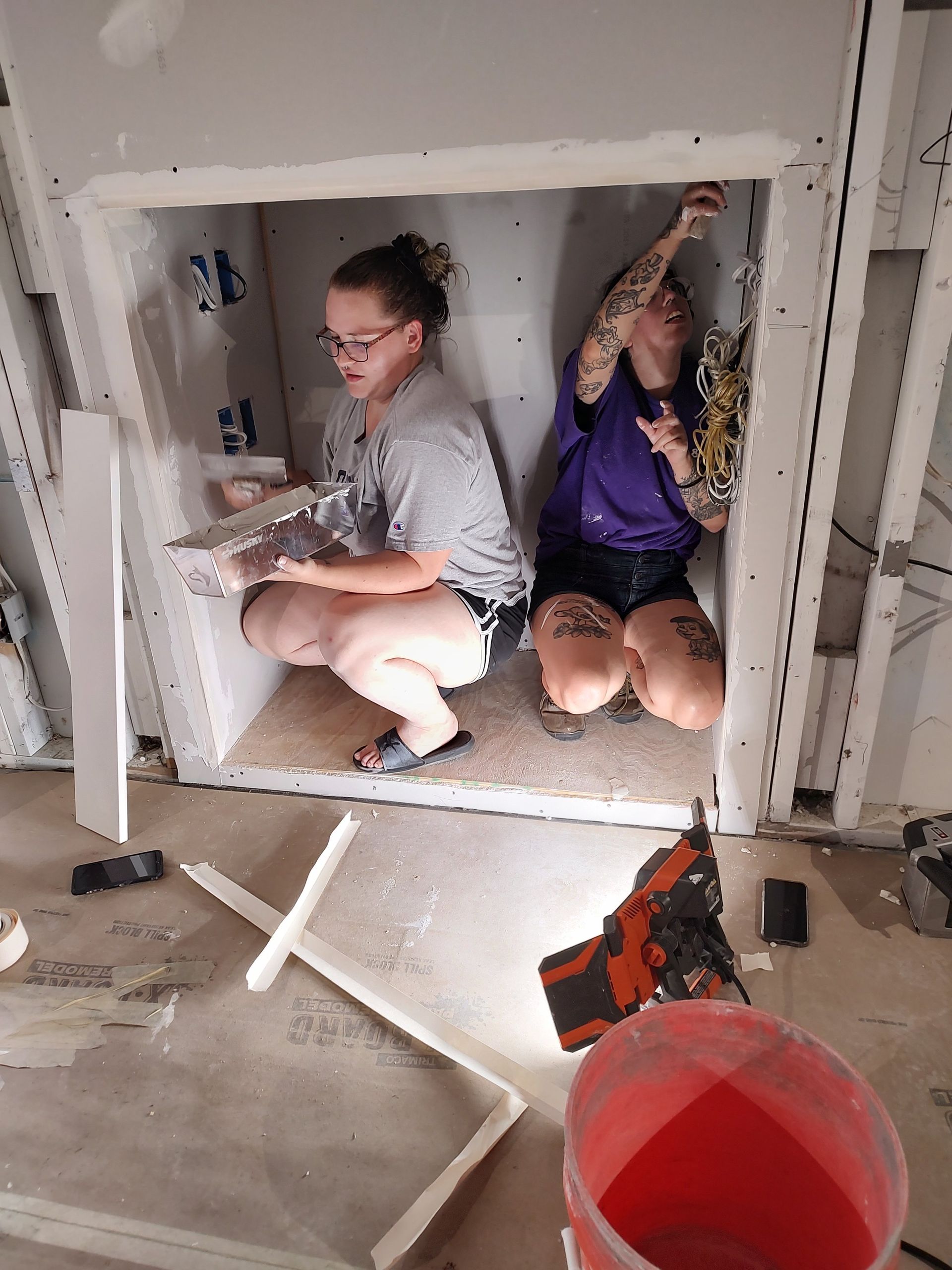 Two people crouch in a wall cavity, applying mud. Interior construction site.
