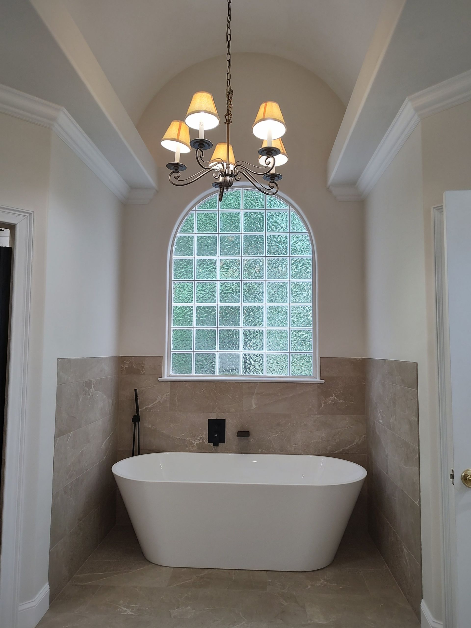 Freestanding white bathtub in a bathroom with arched window, chandelier, and light-colored stone walls.