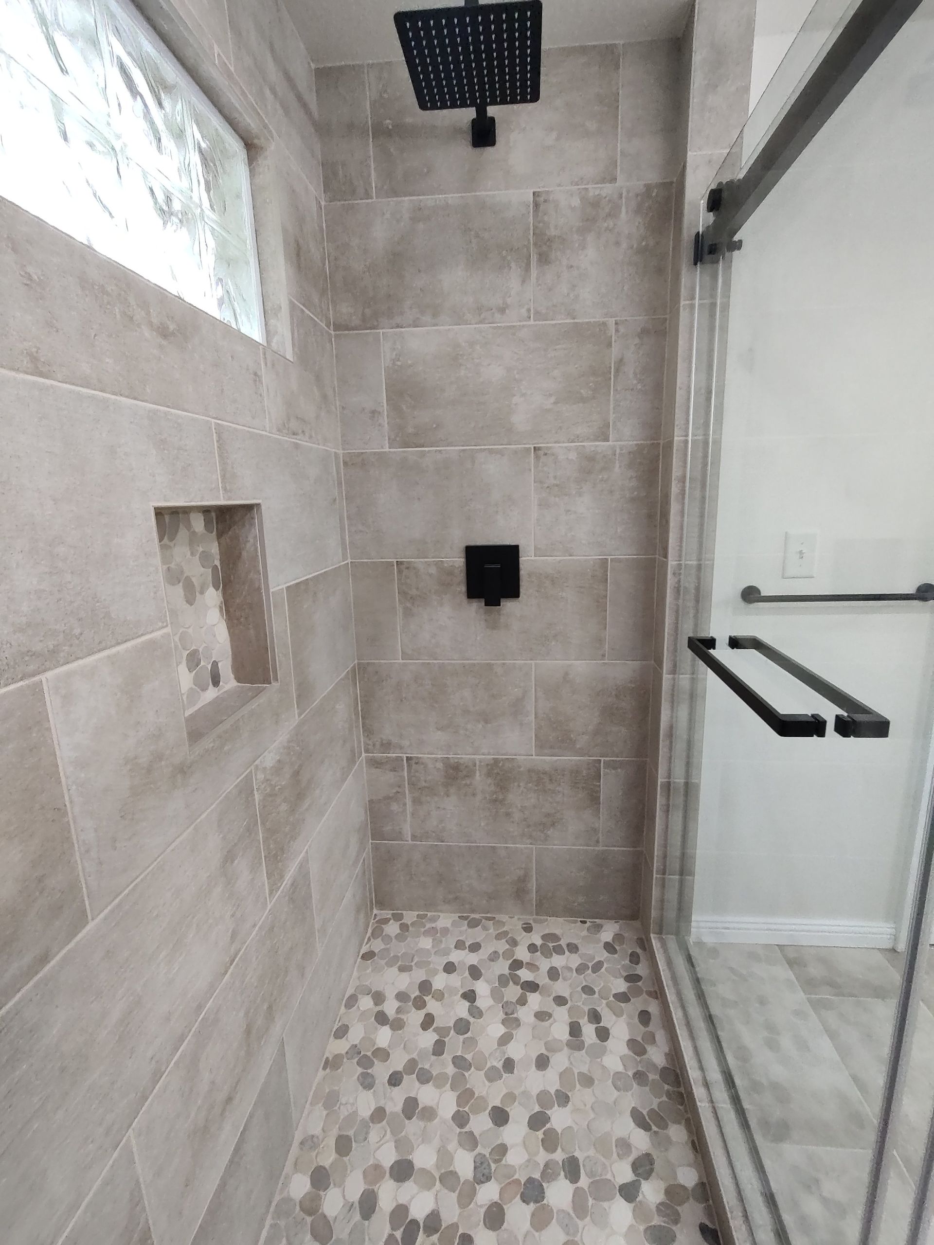 Shower with stone-like tile, pebble floor, black fixtures, and a glass sliding door.