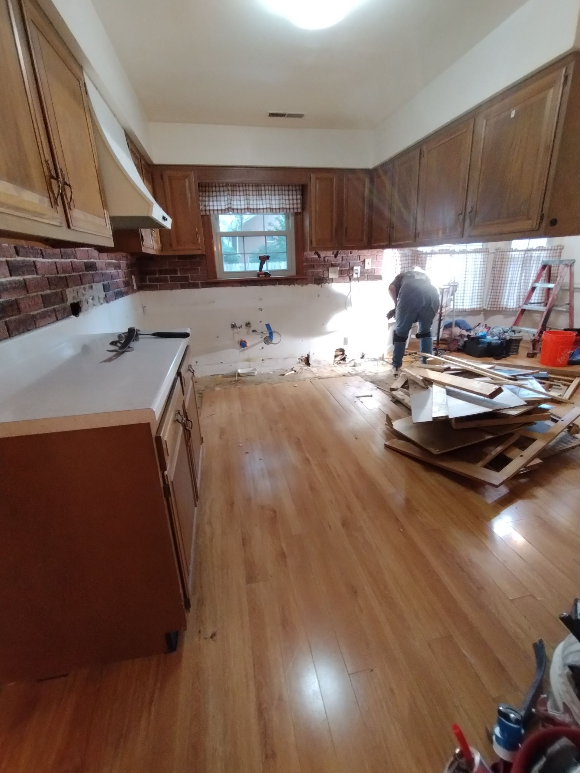 Kitchen renovation: Person working amid debris, cabinets, exposed walls, wood floor, brick backsplash.