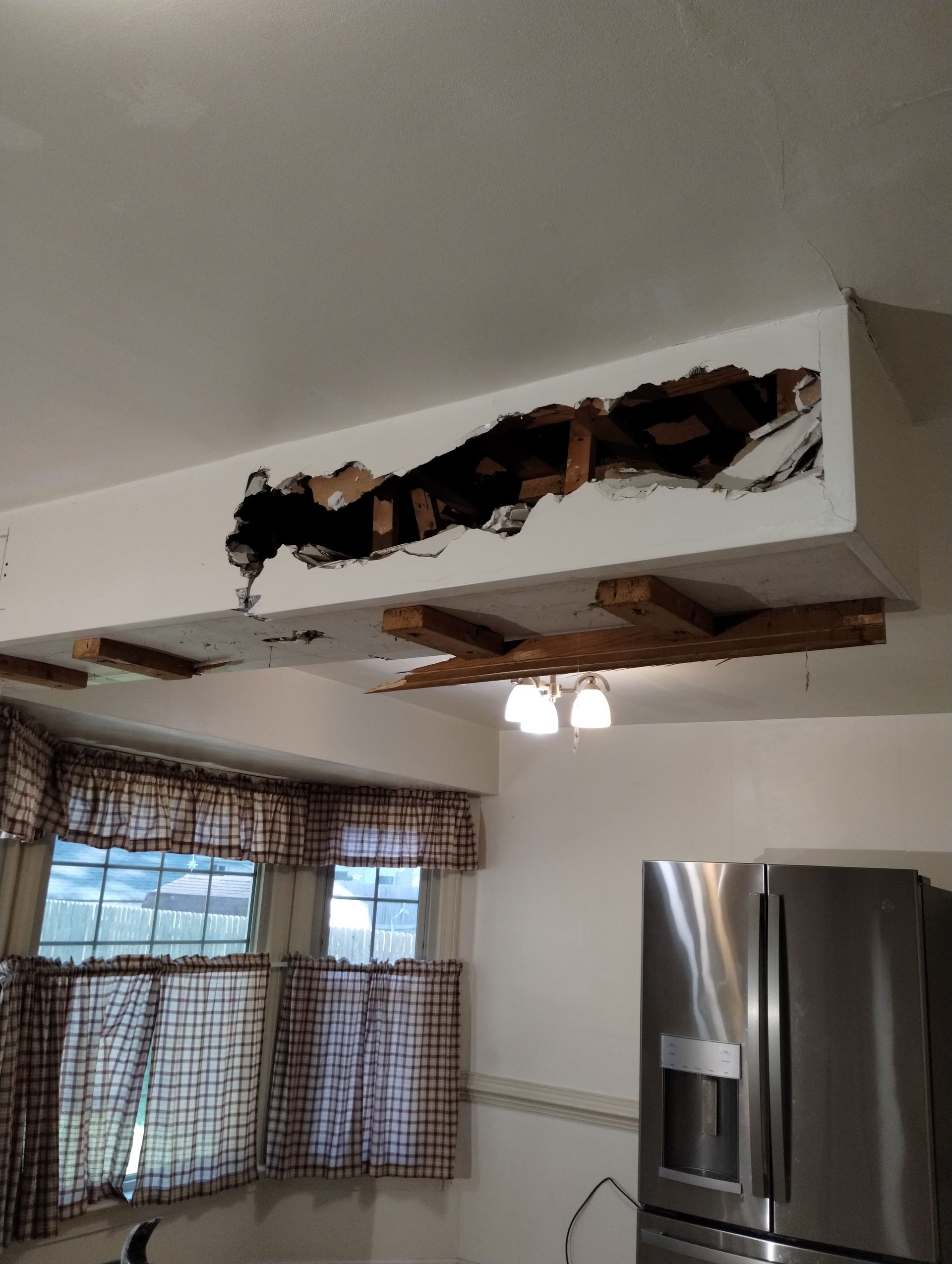 Damage to a ceiling beam revealing wood structure in a kitchen with windows and refrigerator.