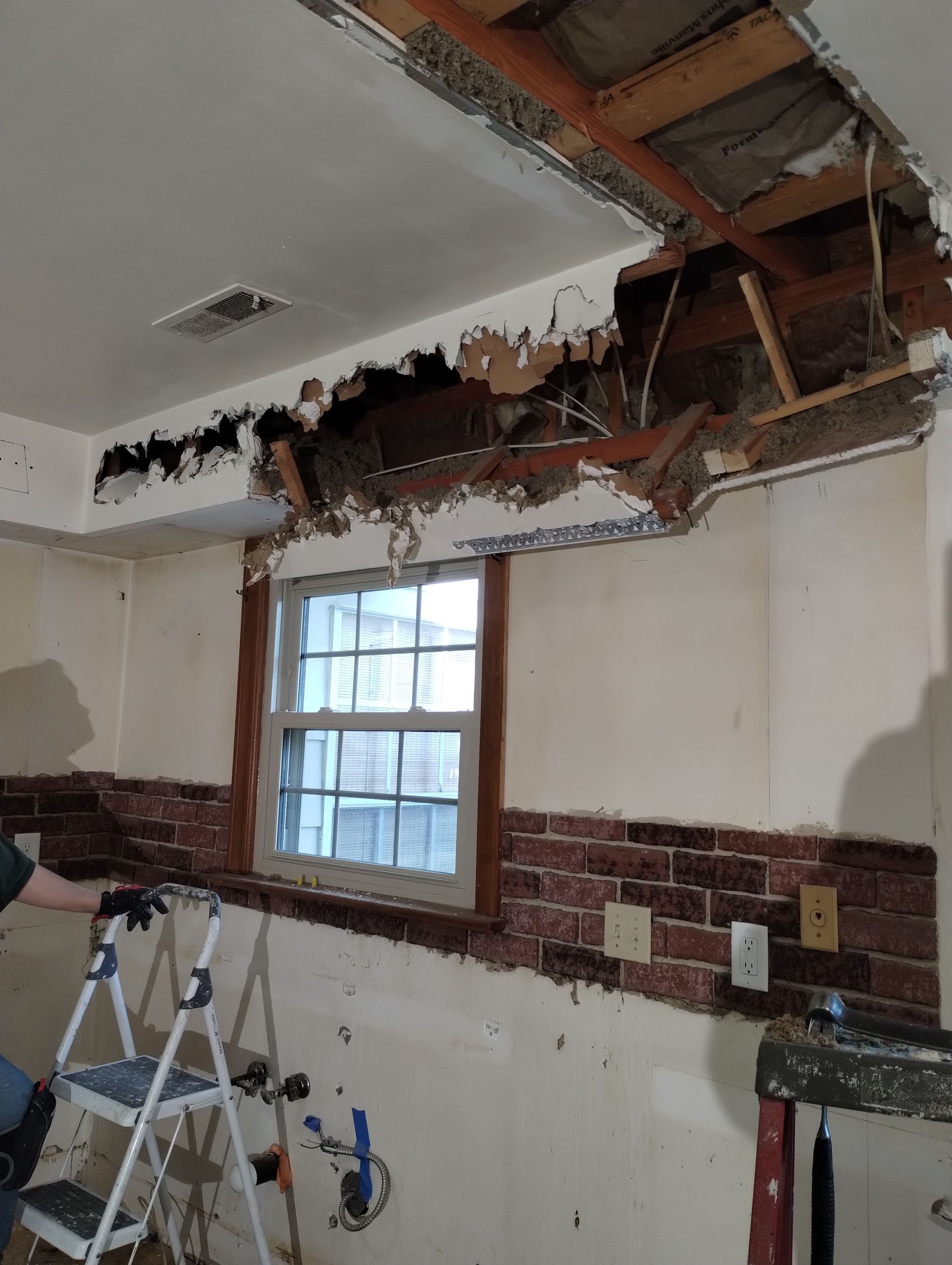 Damaged ceiling and wall reveal wood framing and wires. Window and brick-like trim are visible.