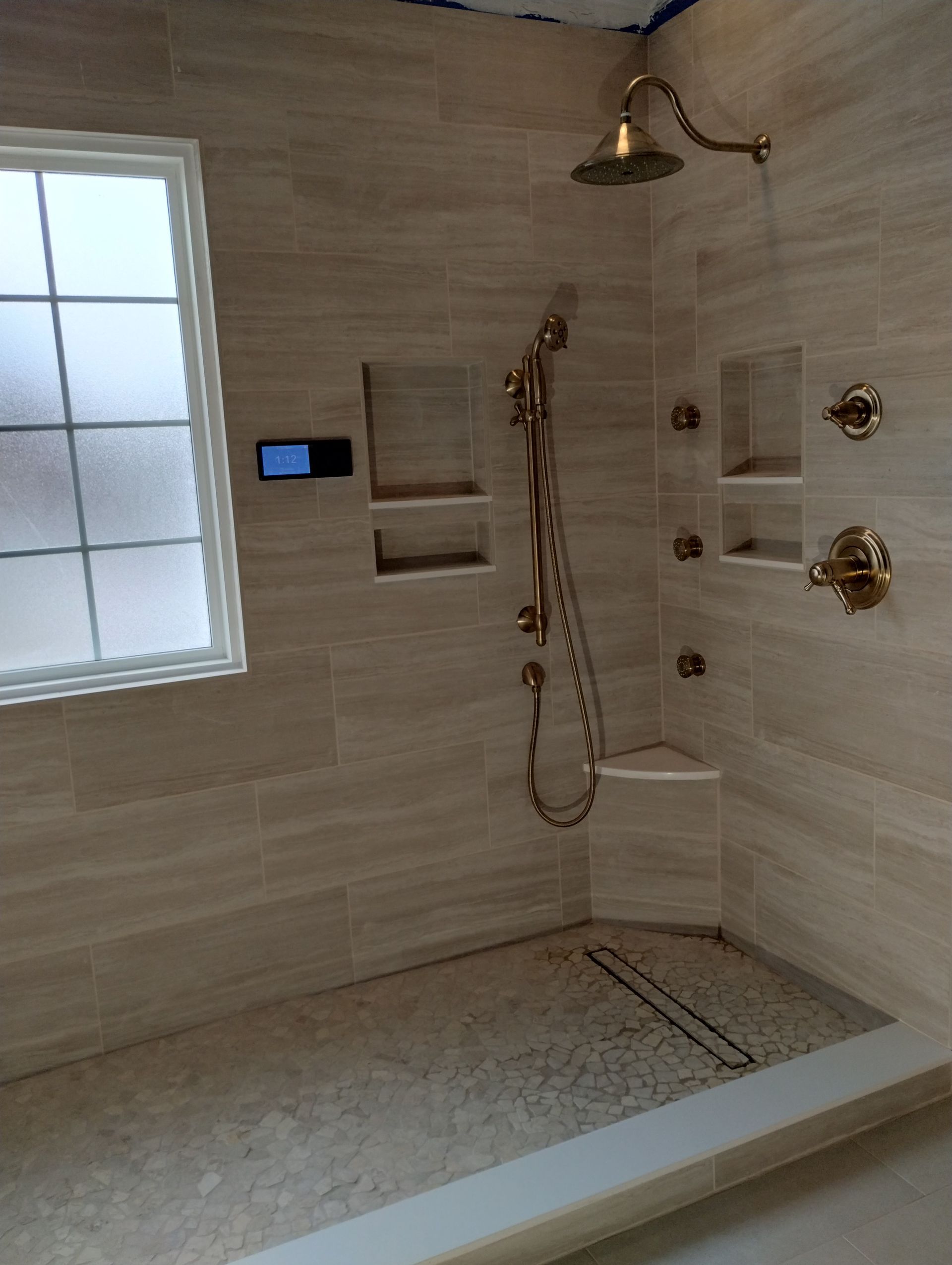 Bathroom shower with beige tile, gold fixtures, a small window, and pebble floor.