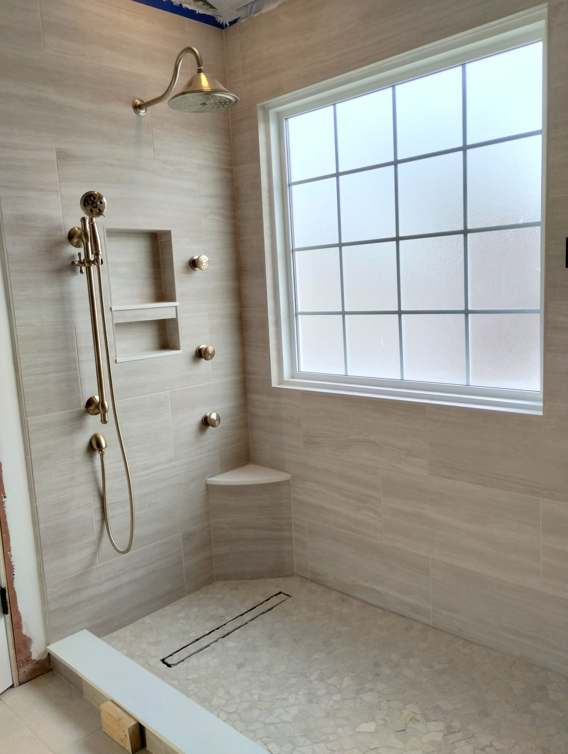 Bathroom with gold shower fixtures, stone tile walls, window with frosted glass, and corner bench.