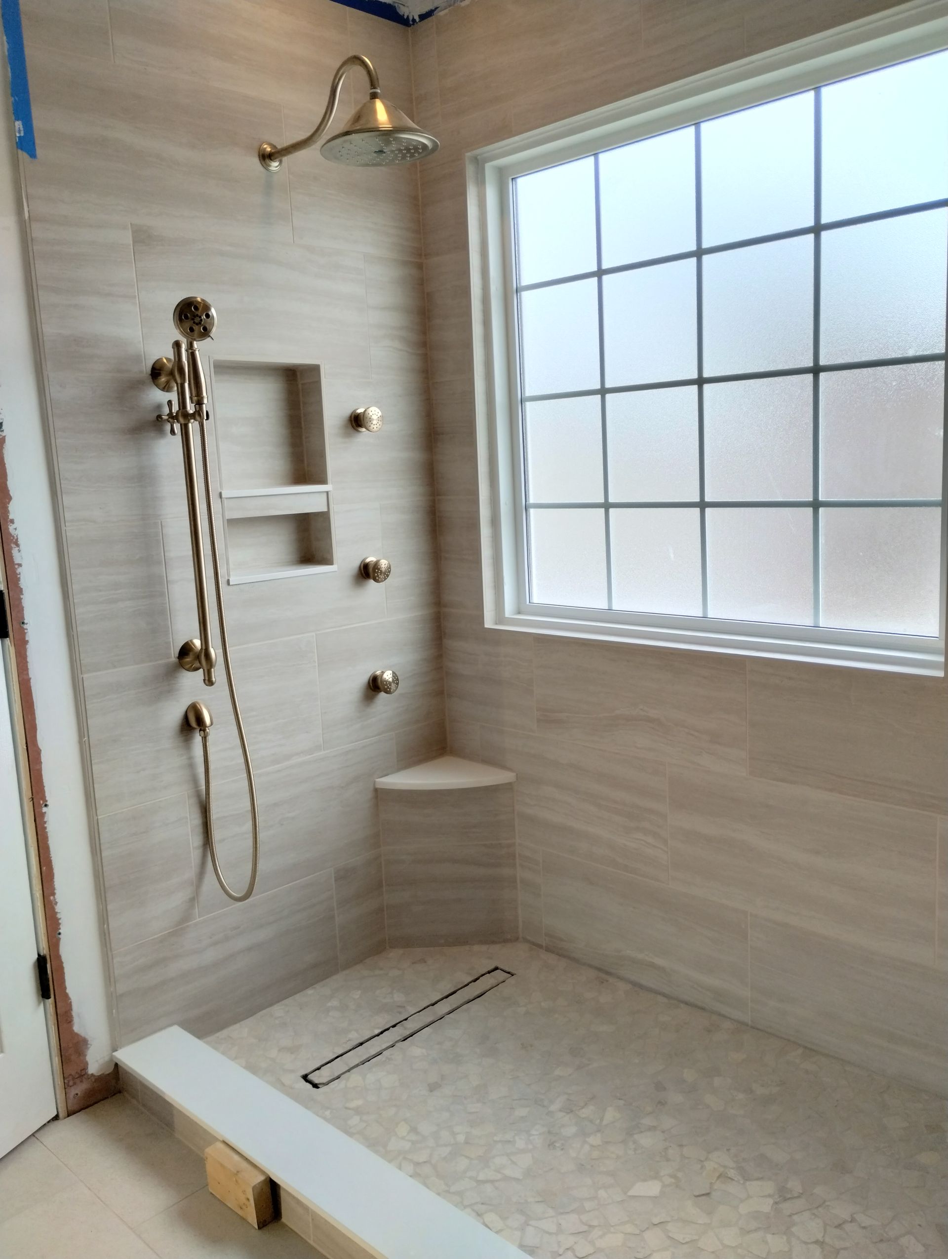 Bathroom shower with gold fixtures, a window, and light-colored tiled walls and floor.