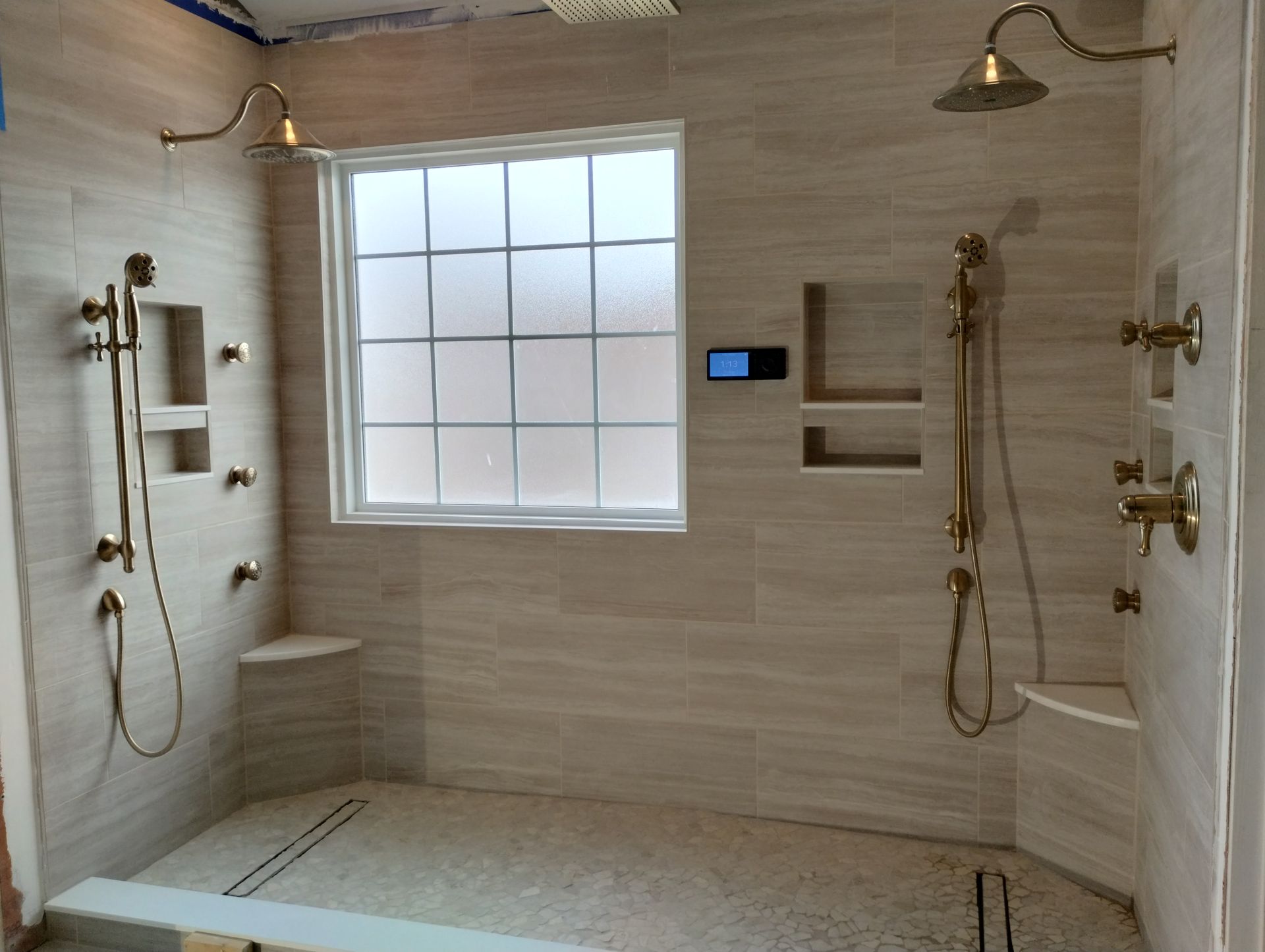 Shower with beige tiles, gold fixtures, and window, featuring multiple showerheads and built-in shelves.