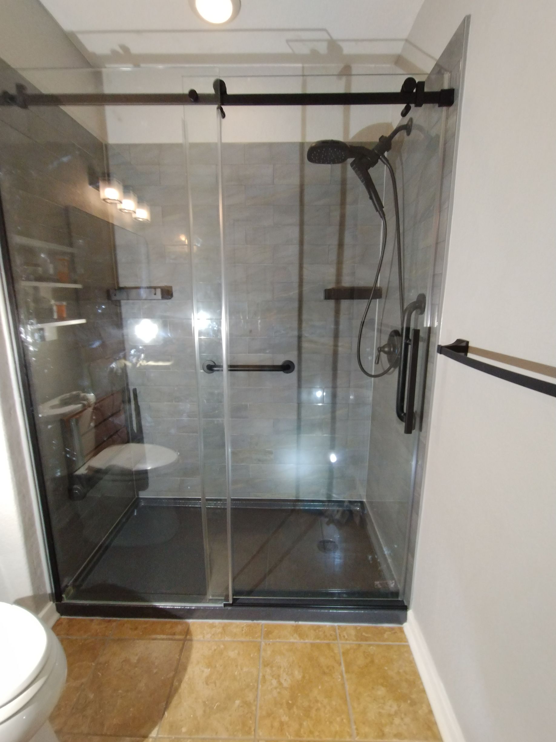 Modern accessible shower with glass doors, dark frame, and grab bars; gray tile walls, seat.