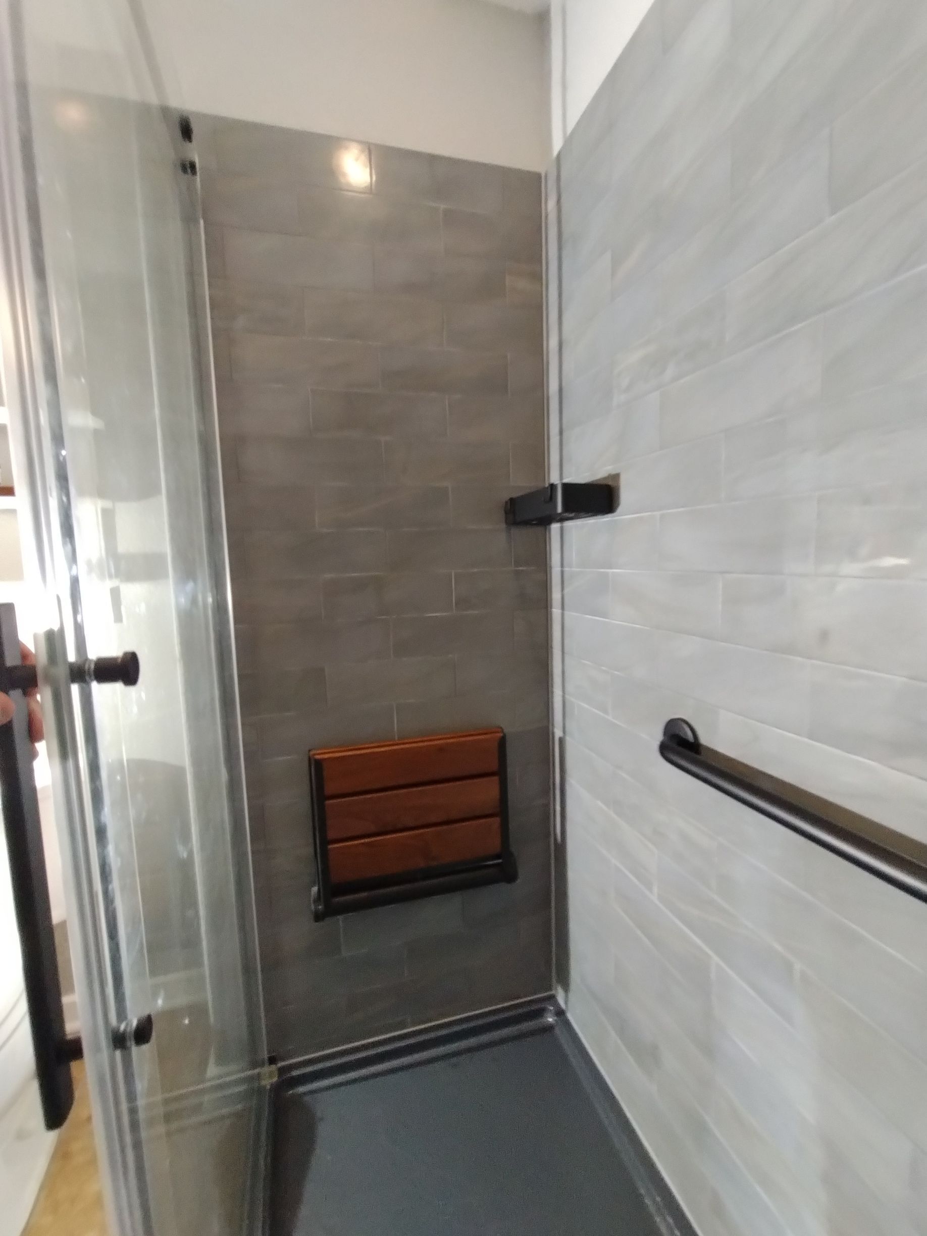 Accessible shower with a fold-down wooden seat, grab bar, and glass door, gray and brown tones.