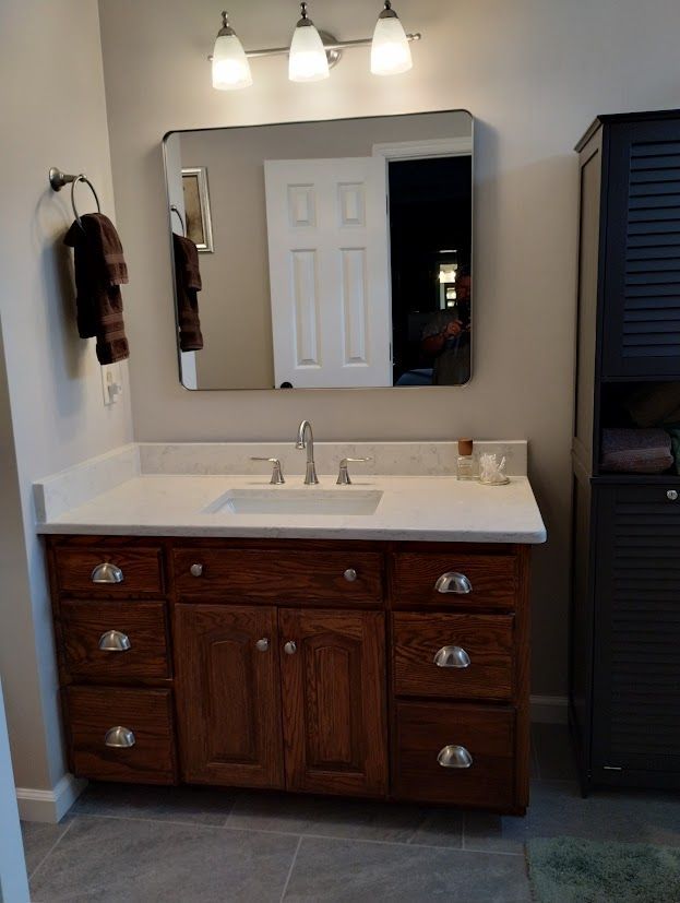 Bathroom vanity with wood cabinets, white countertop, and mirror; dark wood storage cabinet on the right.
