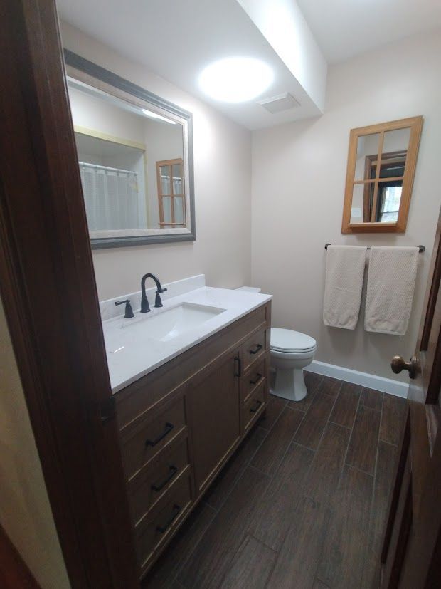 Bathroom with wood-look floor, vanity, toilet, mirror, skylight. Beige walls, brown vanity and door. Two white towels on wall.