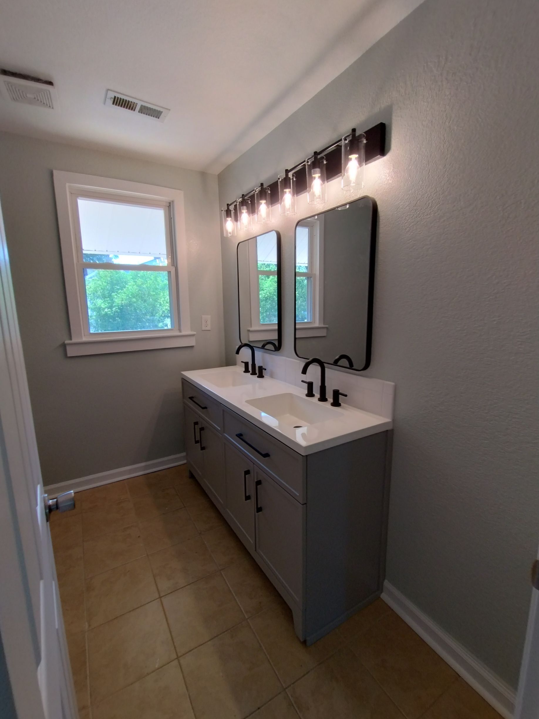 Bathroom with double sink vanity, gray cabinets, black faucets, mirrors, light fixtures, and a window.