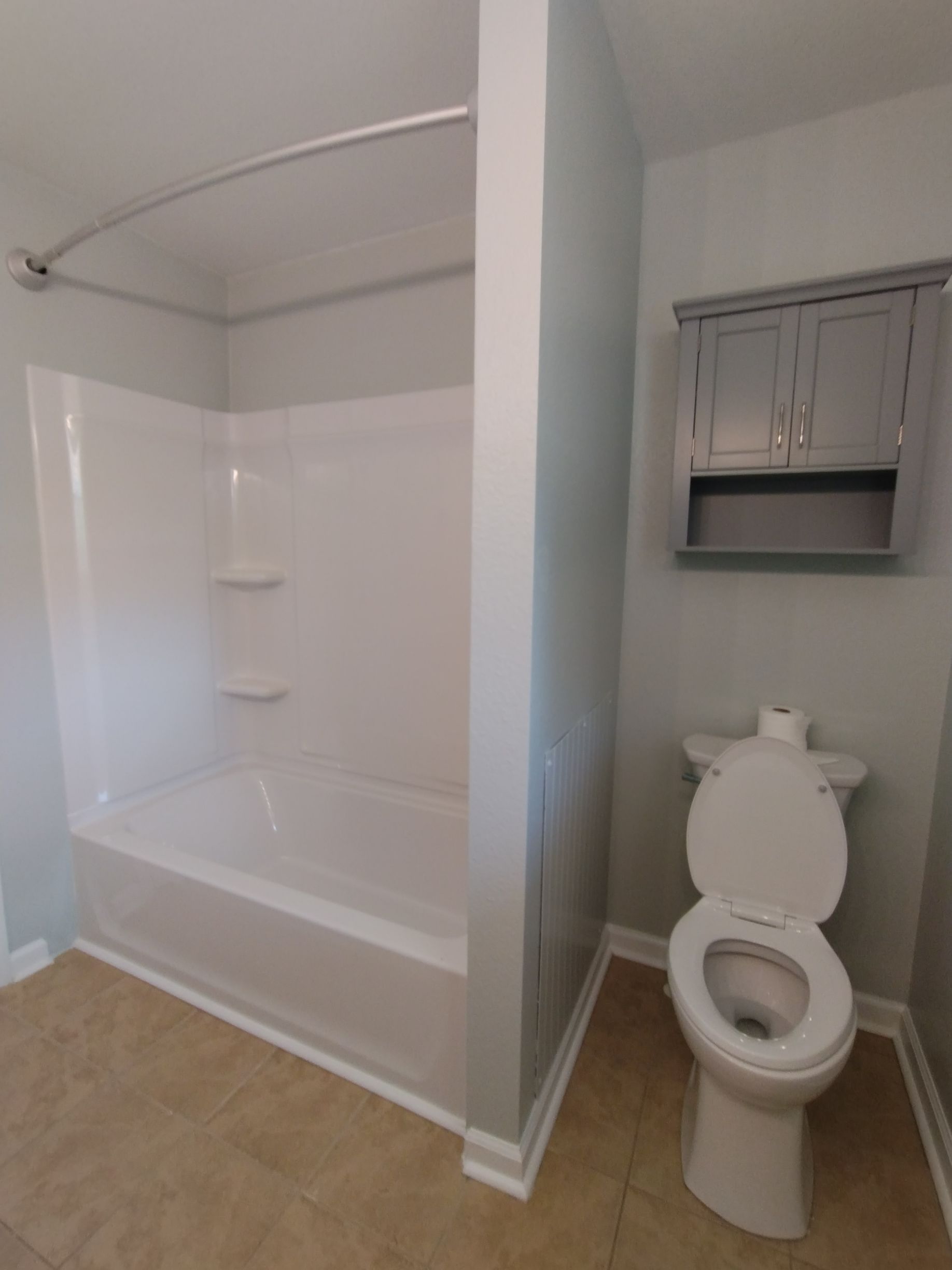 Bathroom with white tub/shower, gray cabinets, and toilet; light blue walls and tan flooring.