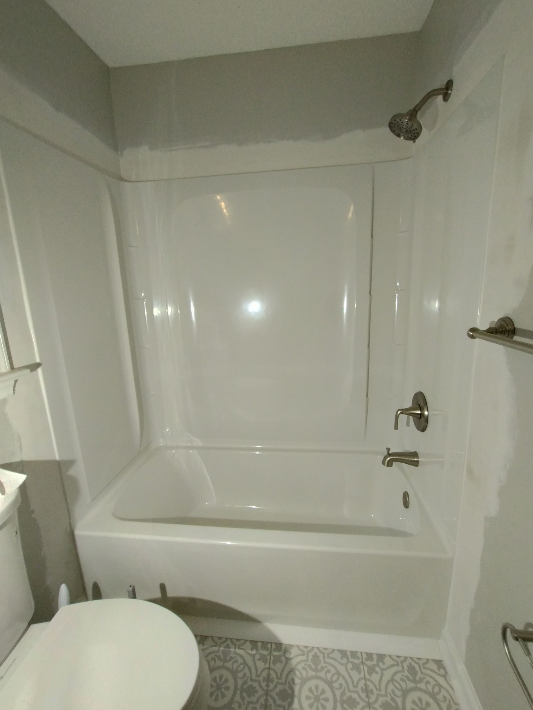 White bathtub and shower enclosure in a bathroom; gray walls, fixtures, and patterned floor.