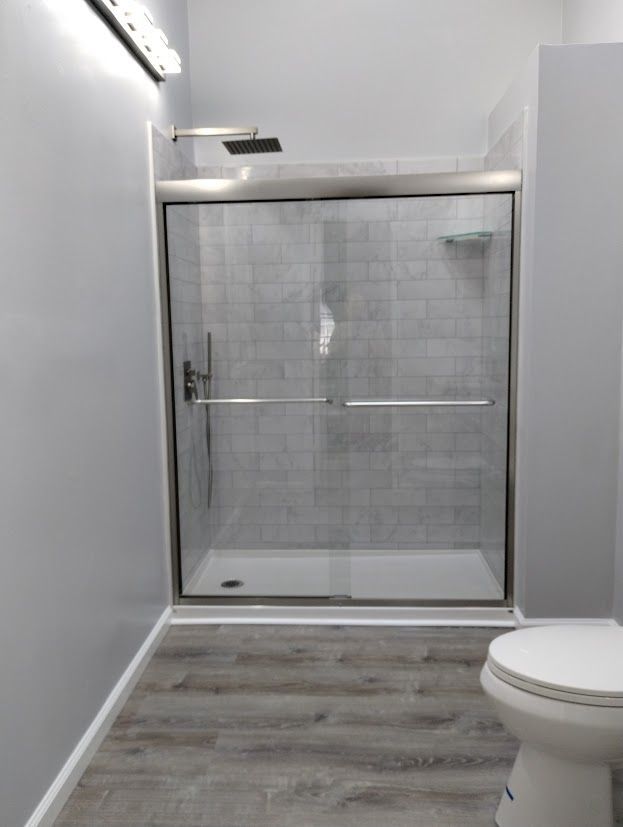 Bathroom with gray walls, tiled shower, glass door, and wood-look floor. Toilet visible on the right.