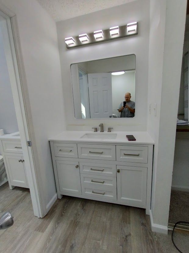 Bathroom with white vanity, large mirror, and light fixtures. A person is reflected in the mirror.