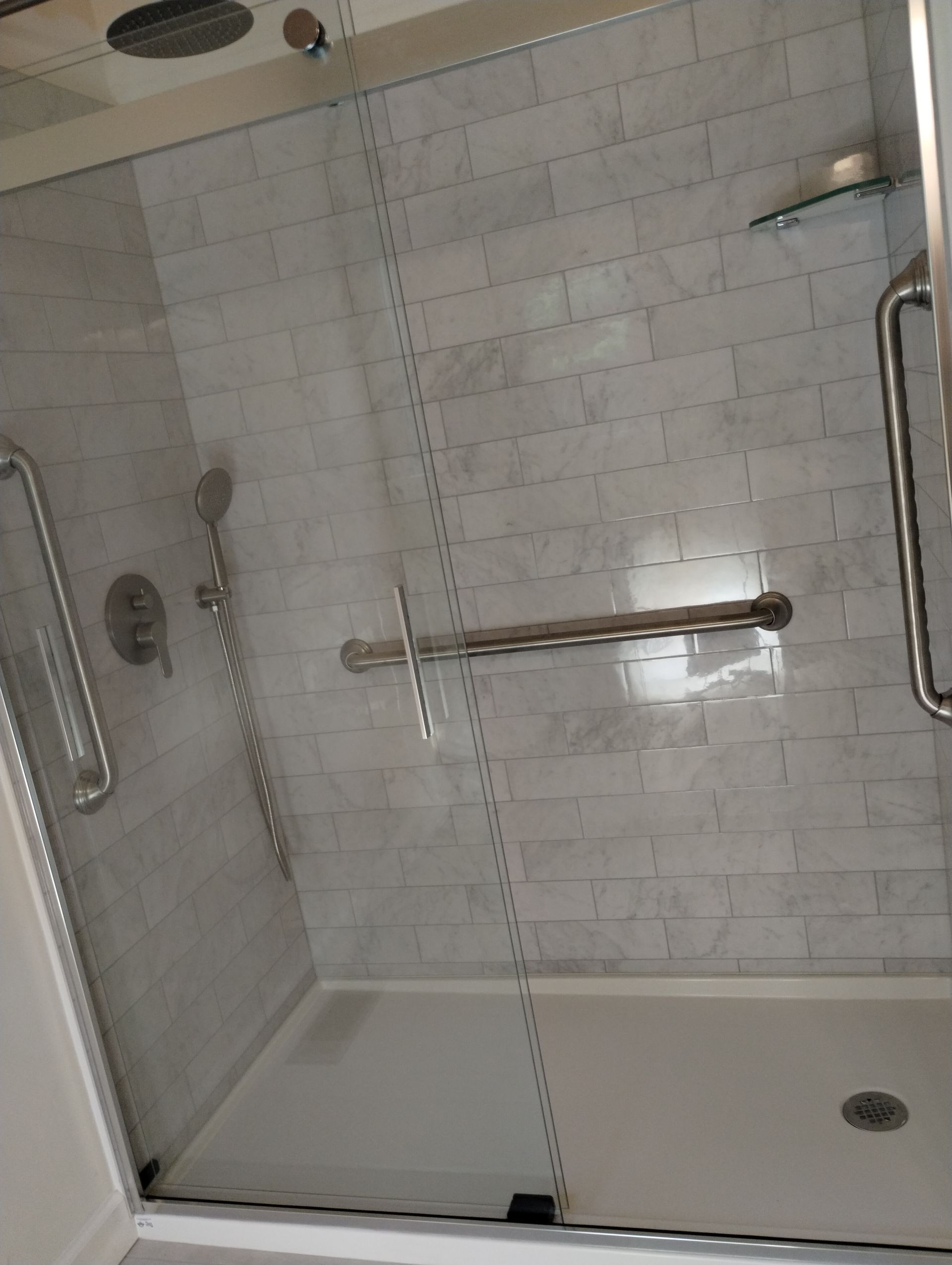 A modern shower with glass doors and light gray brick-patterned tiles, grab bars, and a handheld shower head.