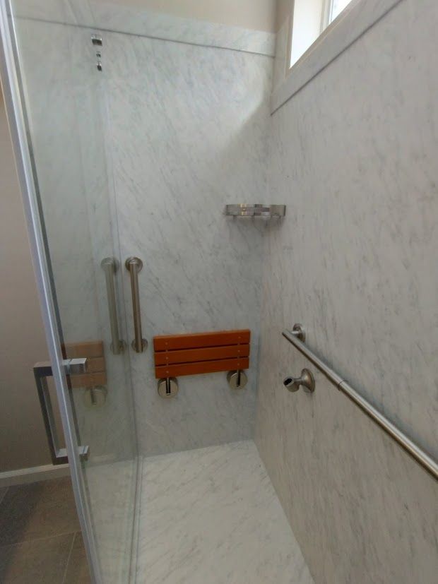 Accessible shower with marble-like walls, glass door, wooden seat, and grab bars.