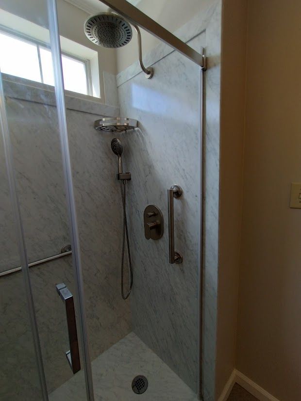 Shower with marble-like walls, glass door, grab bar, and multiple shower heads.