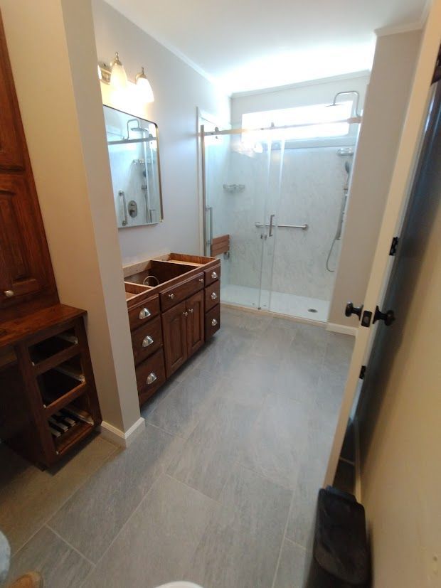 Bathroom renovation: brown vanity and cabinet, glass shower, light gray floor, and beige walls.