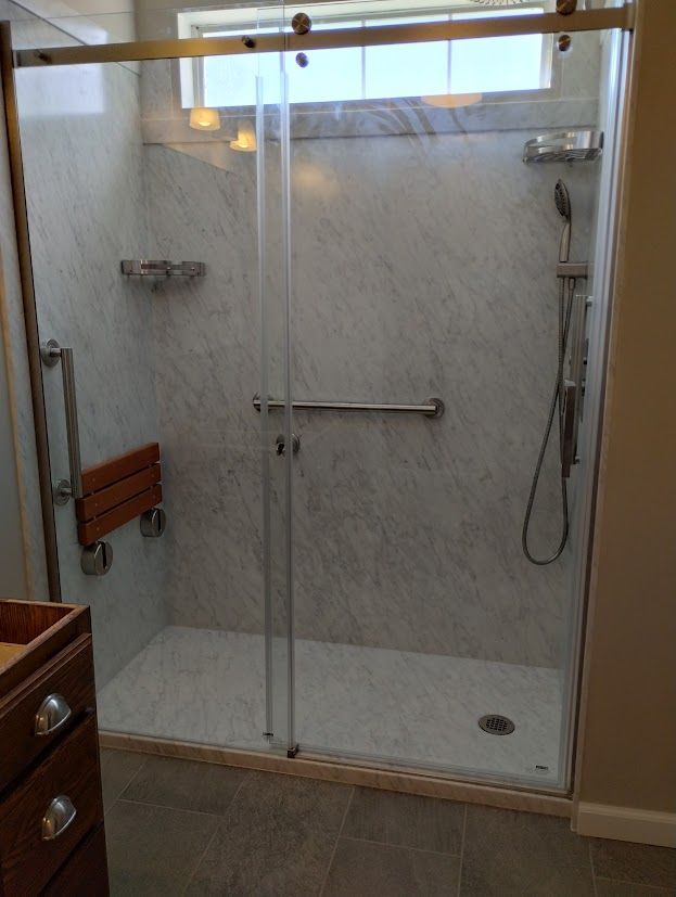 Accessible shower with clear glass doors, marble-like walls, grab bars, a fold-down seat, and hand-held shower head.