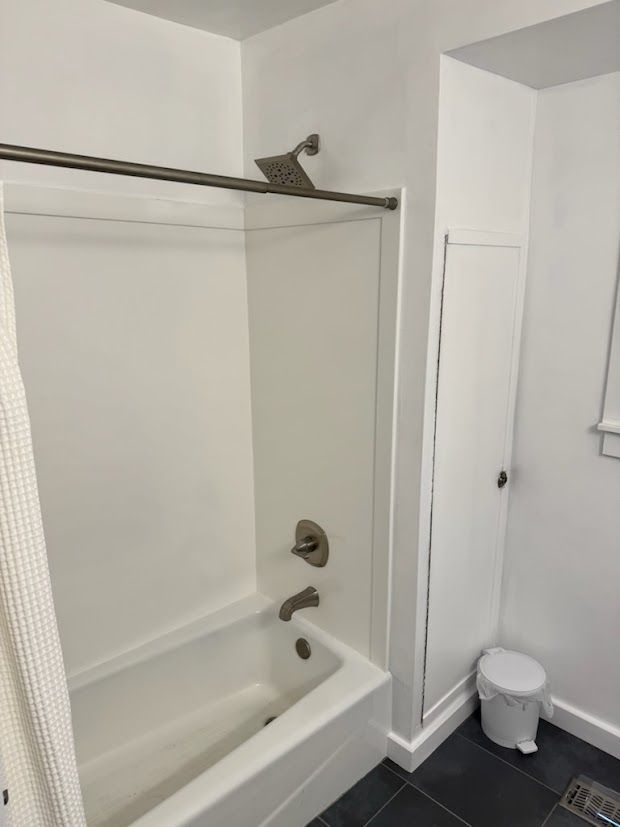 White bathtub and shower with metal fixtures, a narrow closet, and a small trash can.