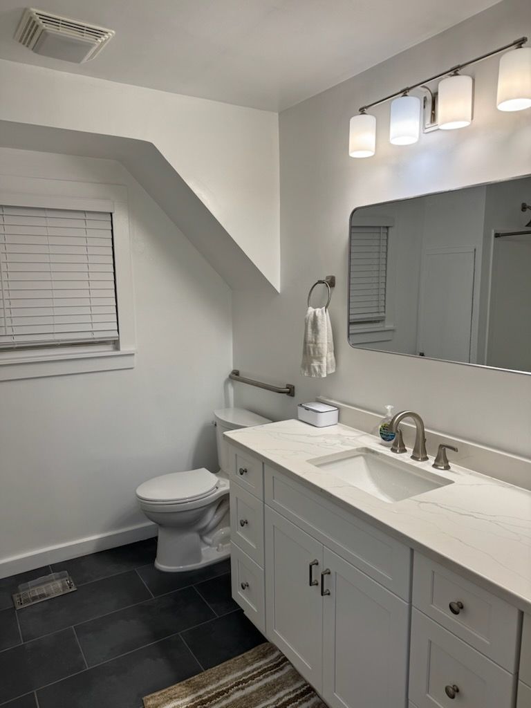 Bathroom with white walls, vanity, and toilet. Black floor. Window with blinds.