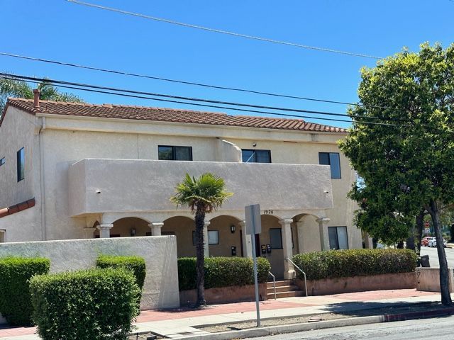 Two-story beige apartment building with red-tiled roof, columns, and shrubs along a sidewalk under a blue sky.