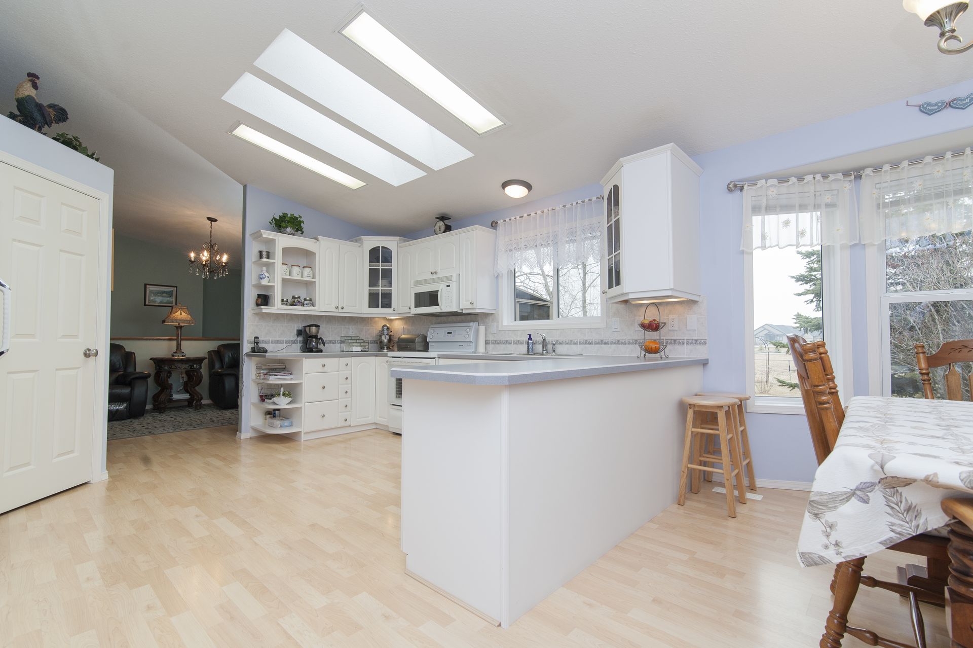 White kitchen with light wood floors, white cabinets, and a blue wall, with a view into a living room.