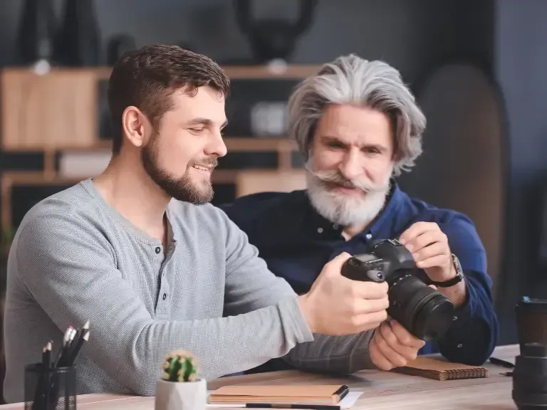 Two men are sitting at a table looking at a camera.