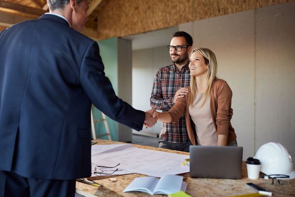 Man in suit shakes hands with woman, couple watches, blueprints on table, interior setting.