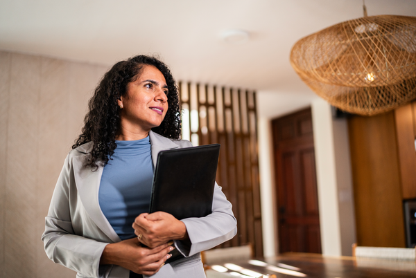 Woman in blazer holds a folder, looking upward in a room with a woven light fixture and wood accents.