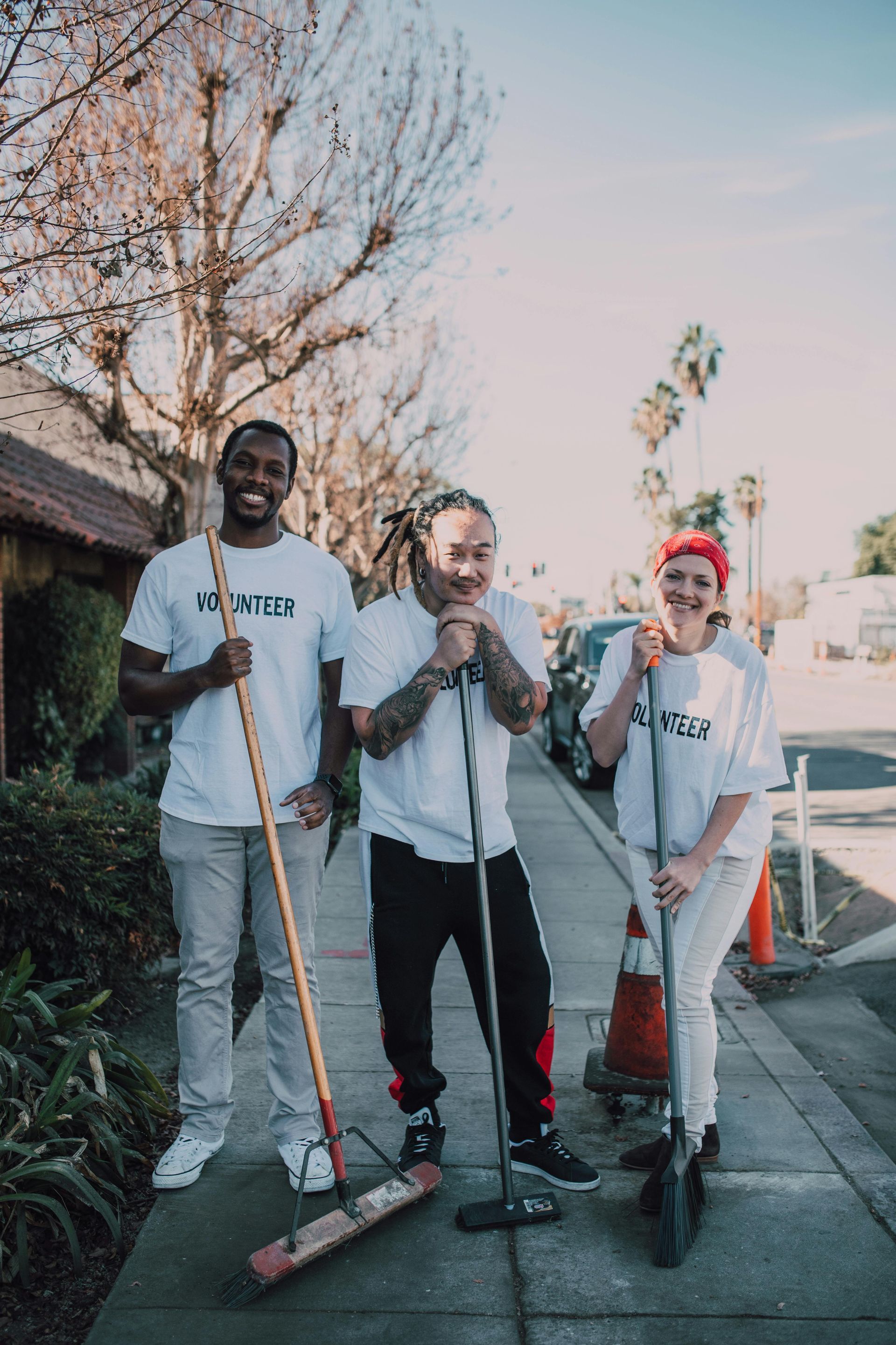 Three people with brooms on a sidewalk, cleaning. One smiles, one rests chin on broom. Sunny day.