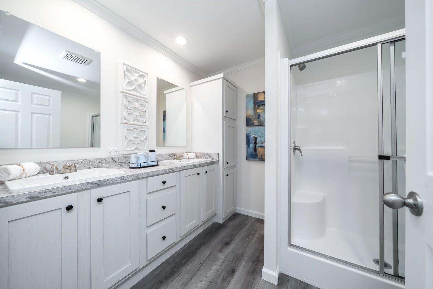 White bathroom with double vanity, walk-in shower, and gray wood-look flooring.