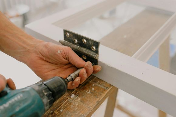 Person using a drill to attach a metal hinge to a white-painted window frame on a wooden surface.