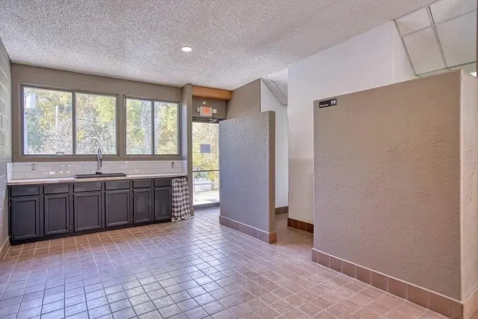 Public restroom with sinks, stalls, and an exit door, featuring windows with outdoor view.