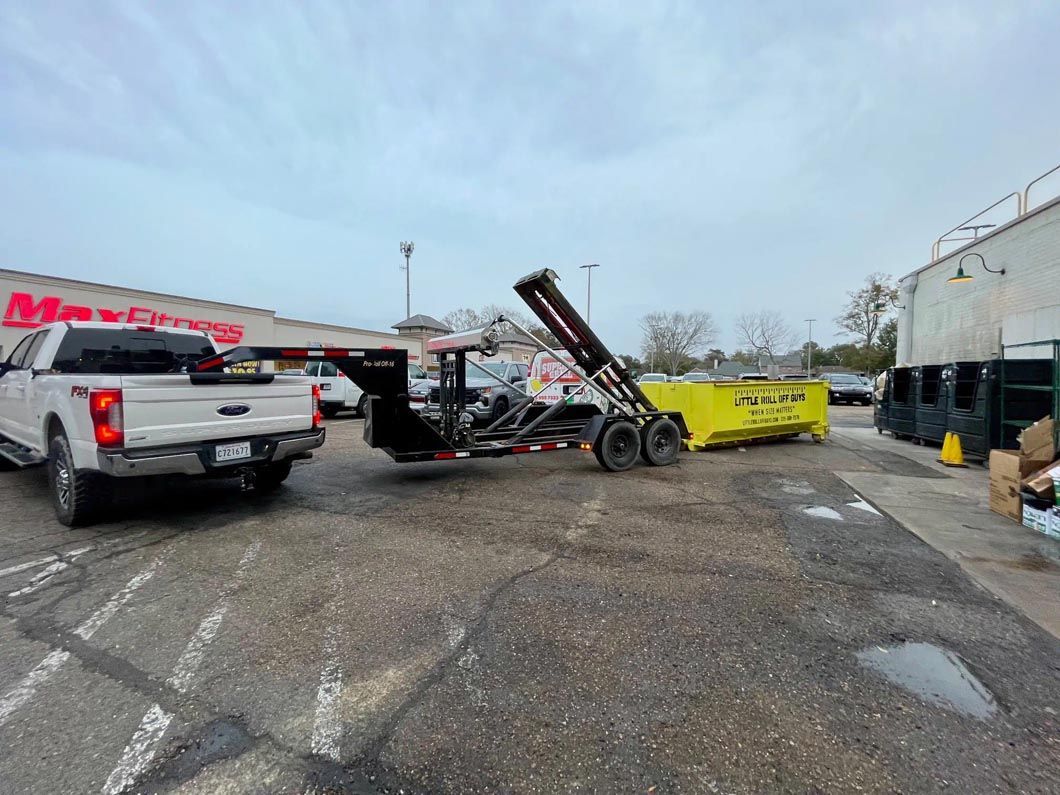 A white truck is towing a yellow dumpster in a parking lot.