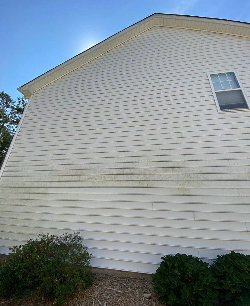 The side of a white house with vinyl siding showing visible discoloration and dirt buildup above the landscaping.