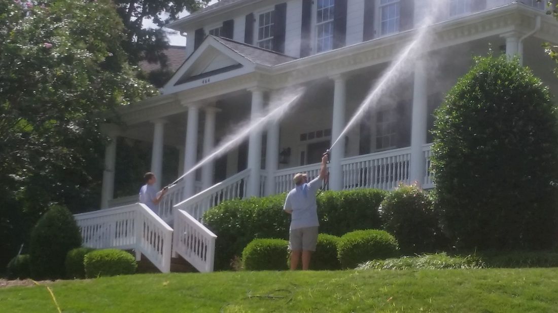 Two people use power washers to clean the white exterior of a two-story house with a front porch.