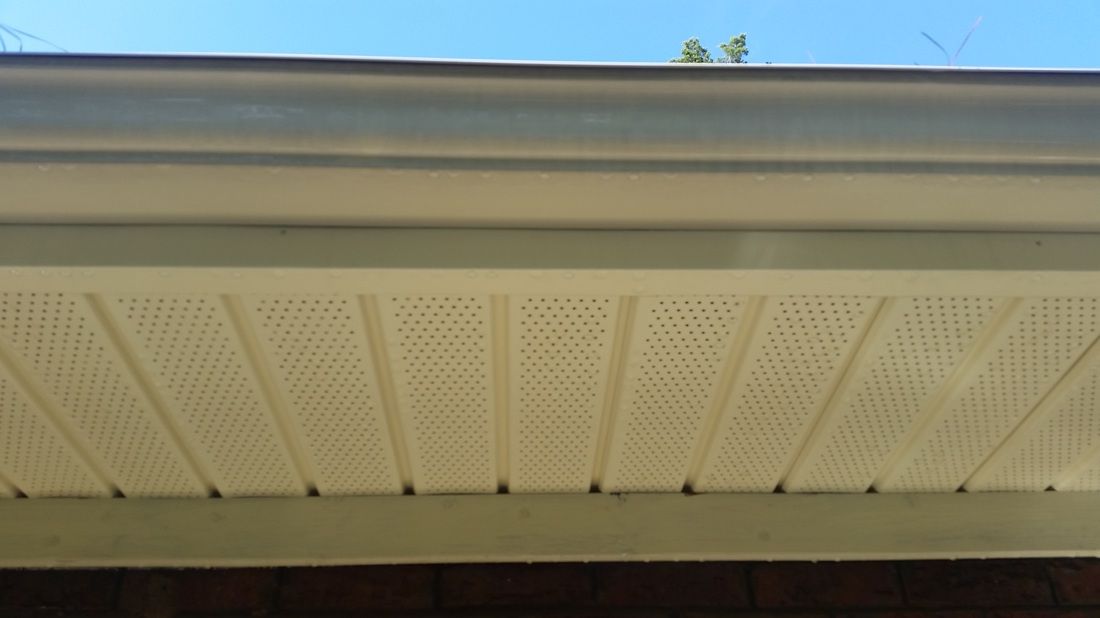 A low-angle view of a white, vented soffit panel beneath a gutter and roofline against a clear blue sky.