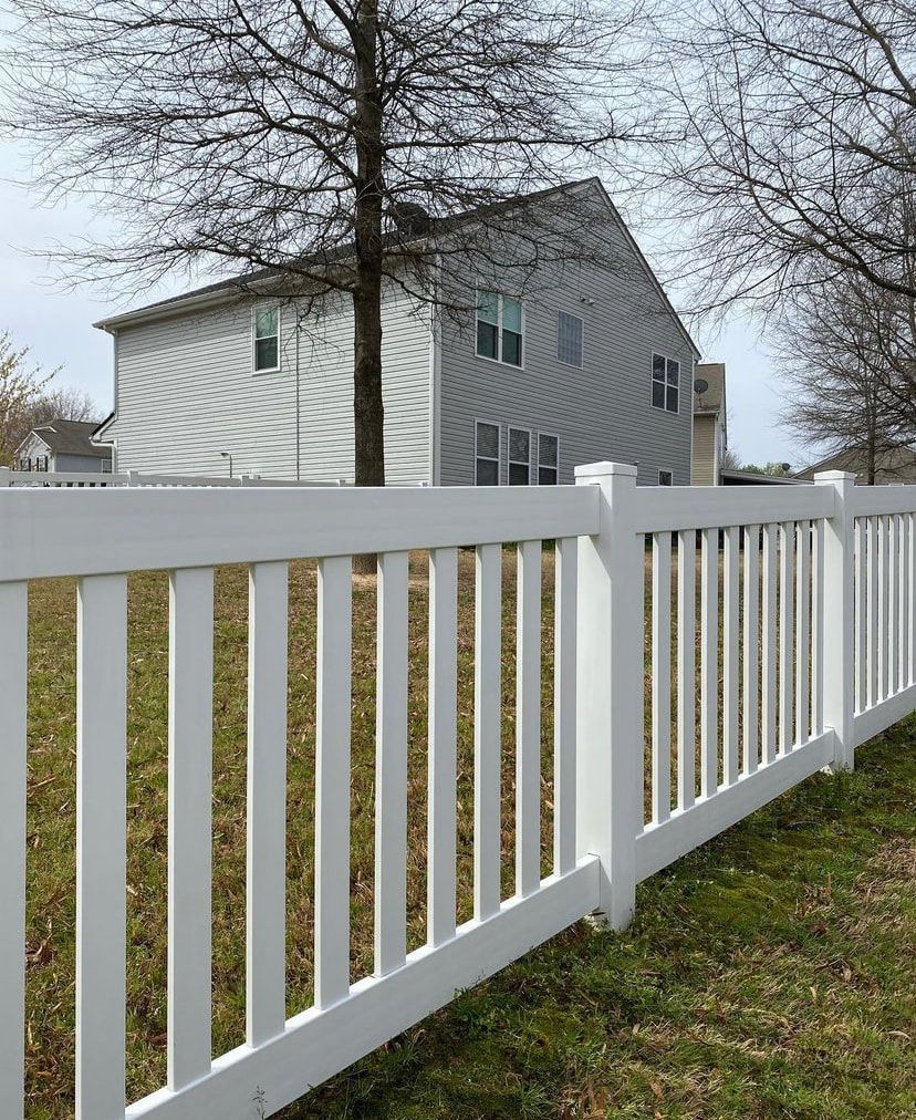 A white vinyl picket fence spans the foreground, with a suburban gray house and leafless trees in the background.