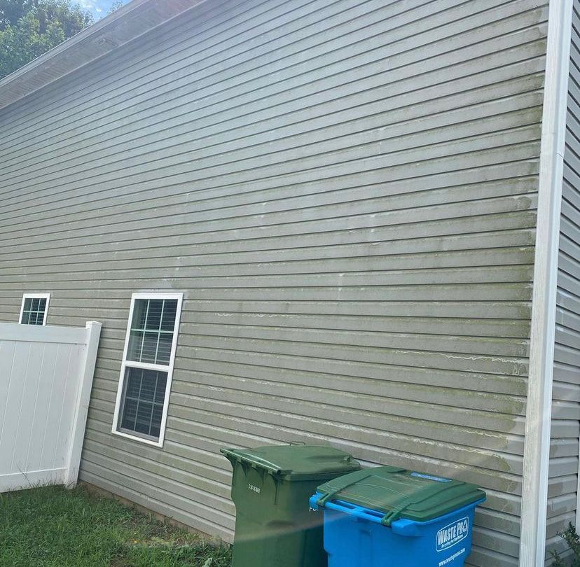 A side view of a house with grey horizontal siding, two windows, and green and blue trash bins outside on the grass.