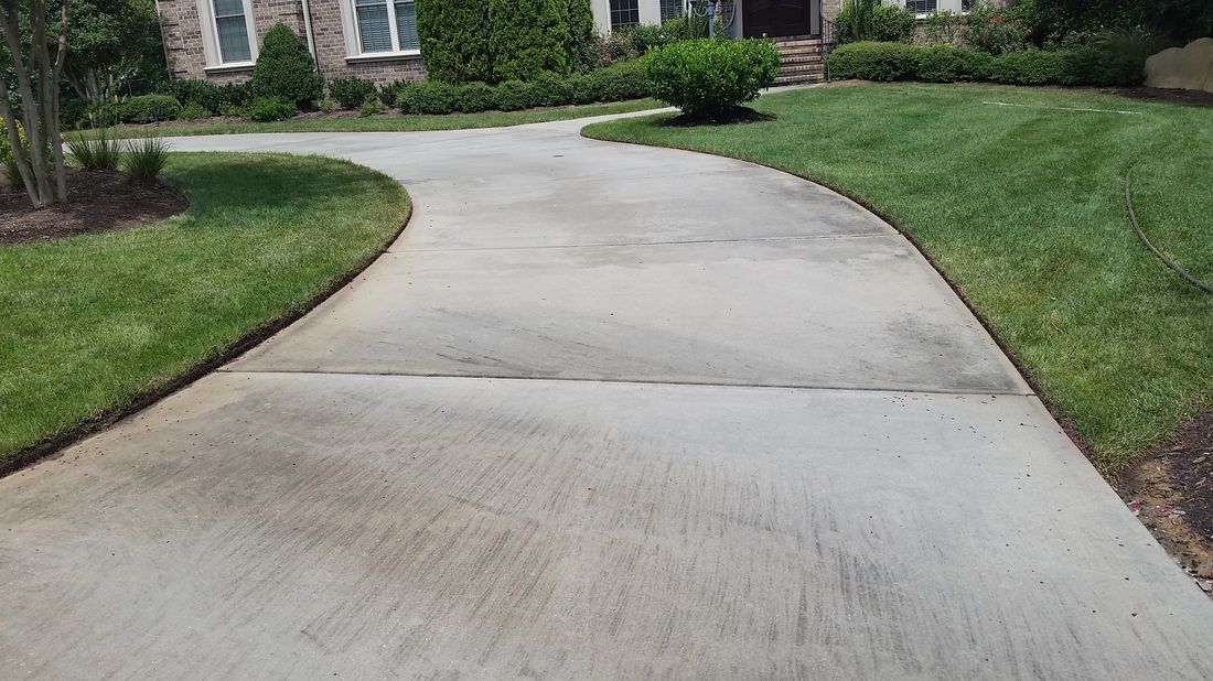 A concrete driveway curves toward a residential house surrounded by a green lawn and landscaping.