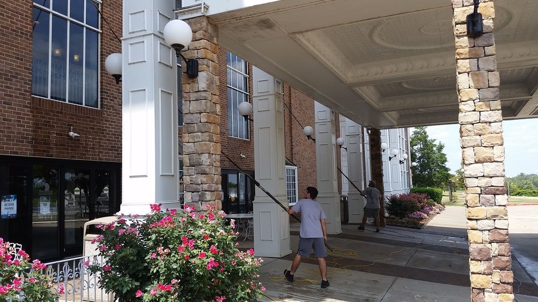 Two people use long-handled tools to clean the exterior stone pillars of a building entrance with a white awning.