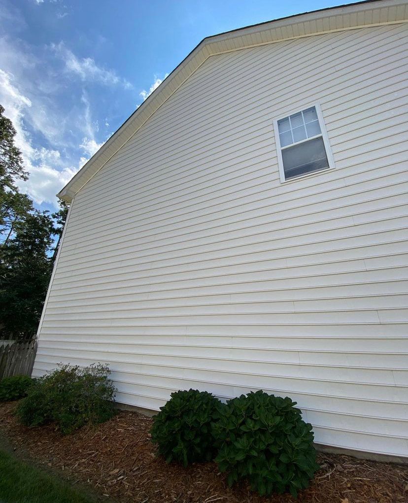 A low-angle view of a white vinyl-sided house exterior with a single window and two round green bushes in front.