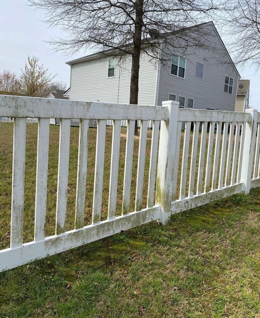 A white picket fence with visible green mildew growth stands in front of a house on a cloudy day.