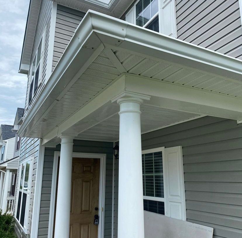 The front porch of a gray-sided home features white columns, a wooden door, and white gutters.