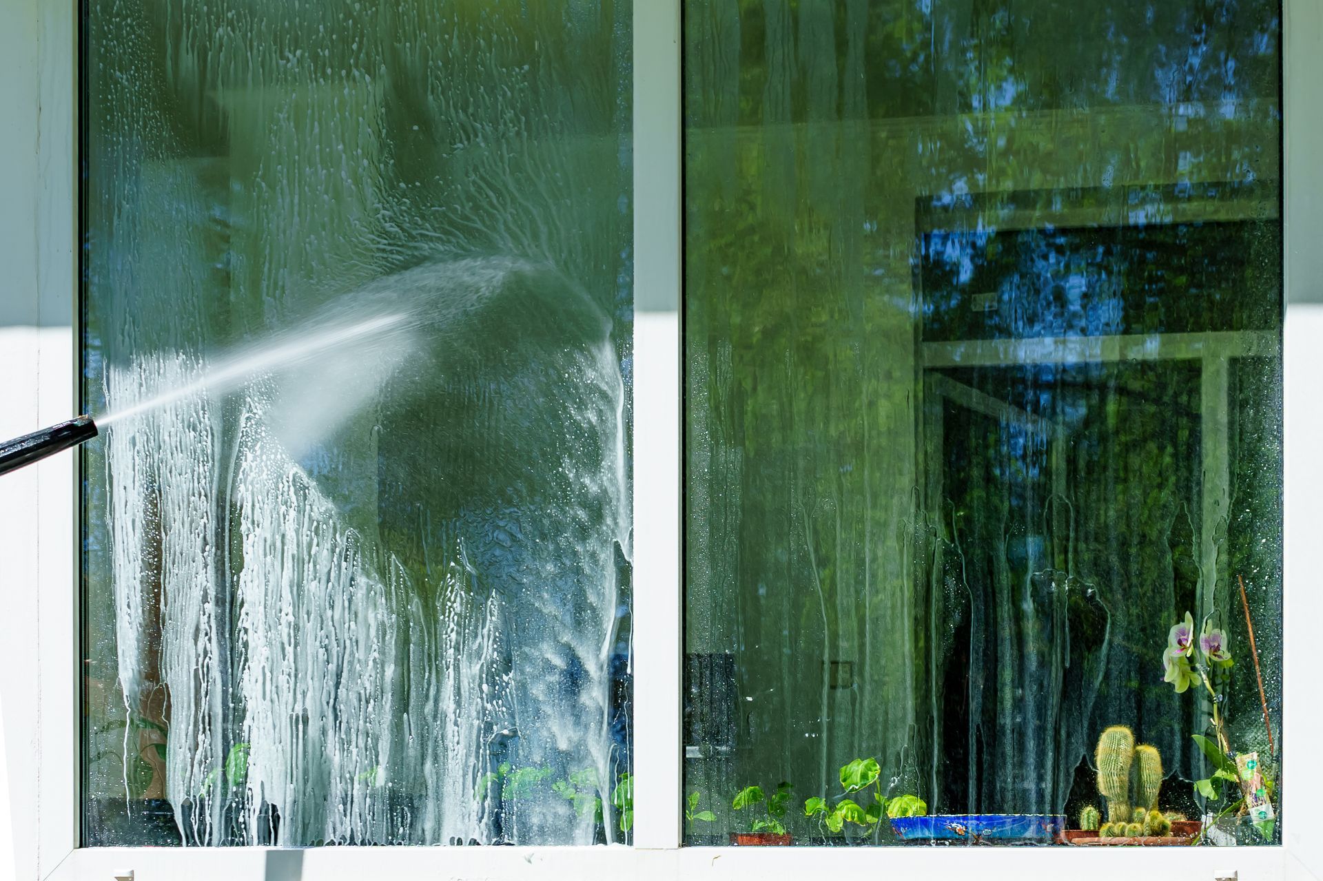 A pressure washer sprays a stream of water onto a glass window, cleaning a dirty pane next to one that is already clear.