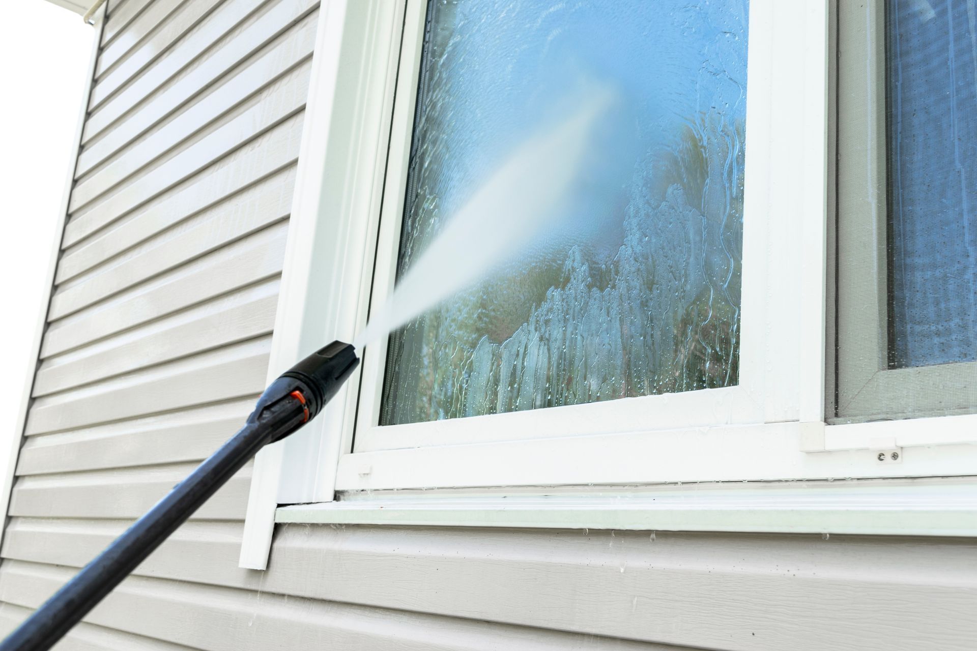 A close-up view of a power washer spraying water onto a window of a house with light-colored vinyl siding.