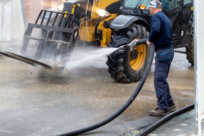 A person in work clothes uses a high-pressure hose to wash the wheel of a yellow construction vehicle.