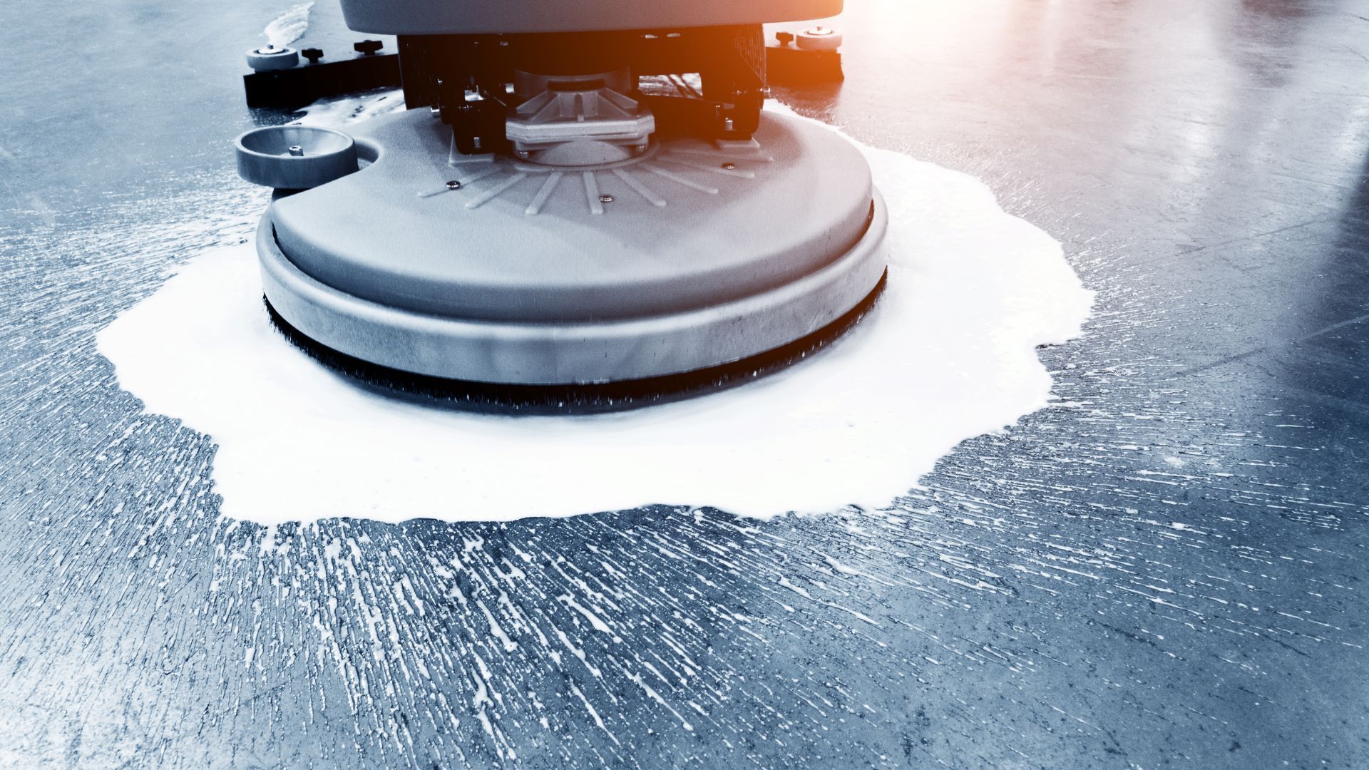 Floor cleaning machine dispensing cleaning solution on a wet, gray floor.
